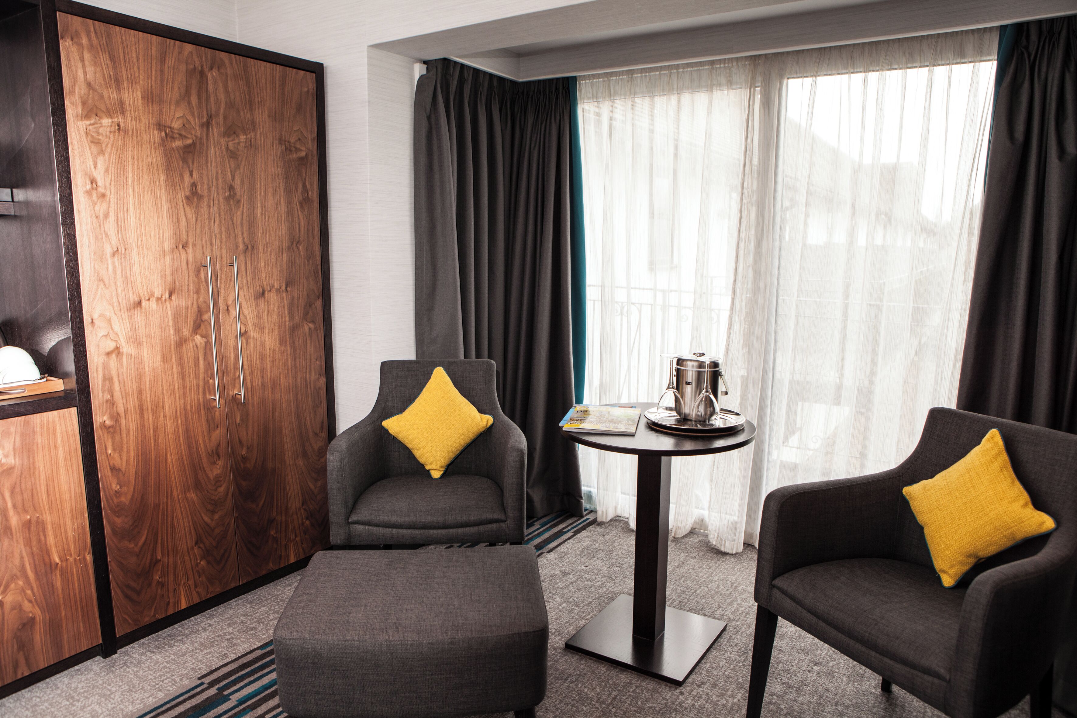 A hotel room with a wardrobe, two armchairs with yellow pillows, a small table with a tray, and a window with sheer curtains.