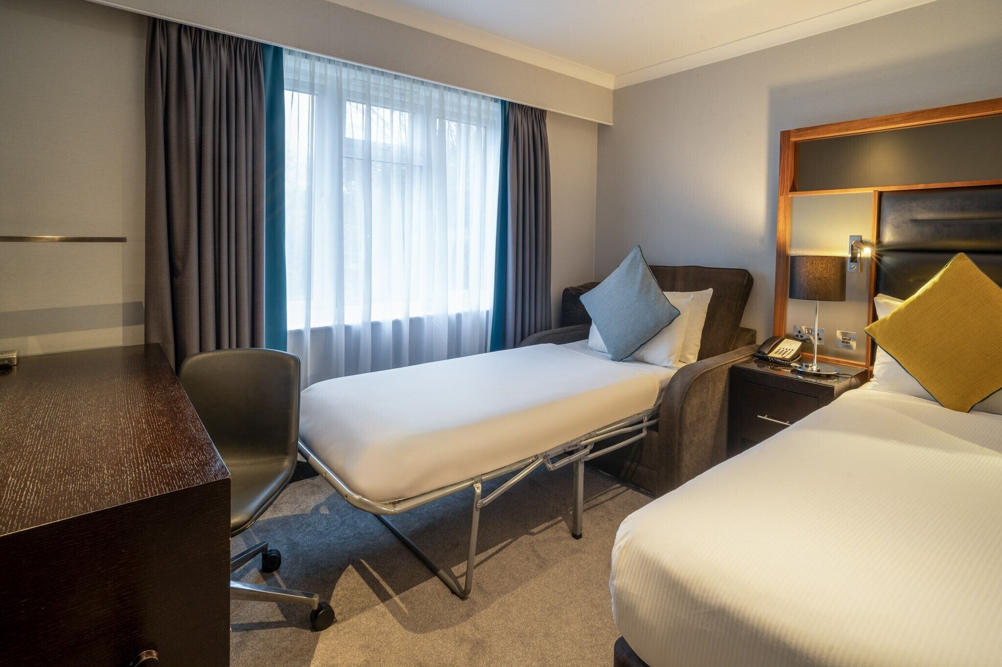 A hotel room with a bed, bedside table, lamp, and a desk with a chair.
