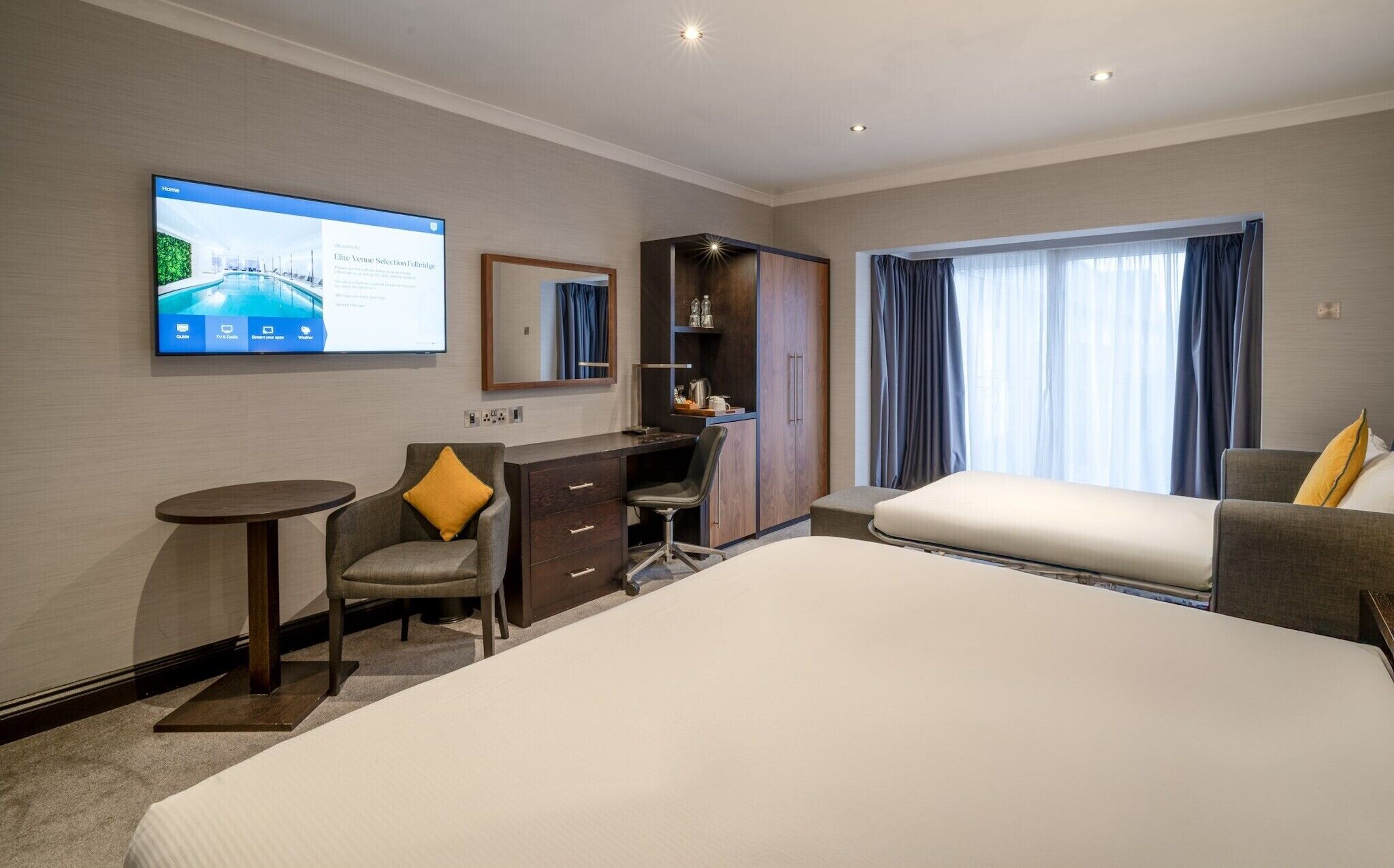 A hotel room with a large bed, a desk with a chair, a TV mounted on the wall, and a window with curtains.
