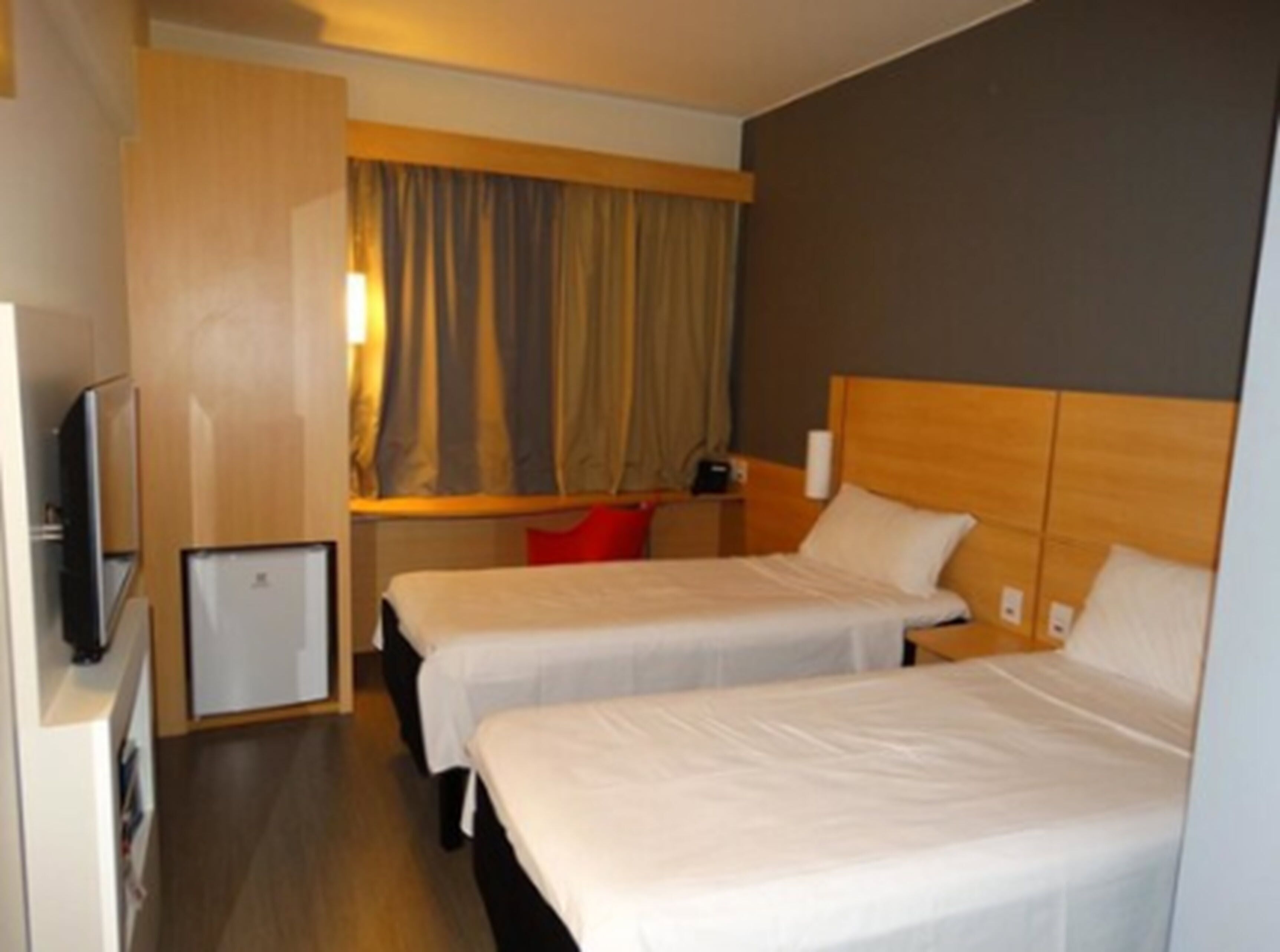 A hotel room with two beds, a small desk, a television, and a window with curtains.