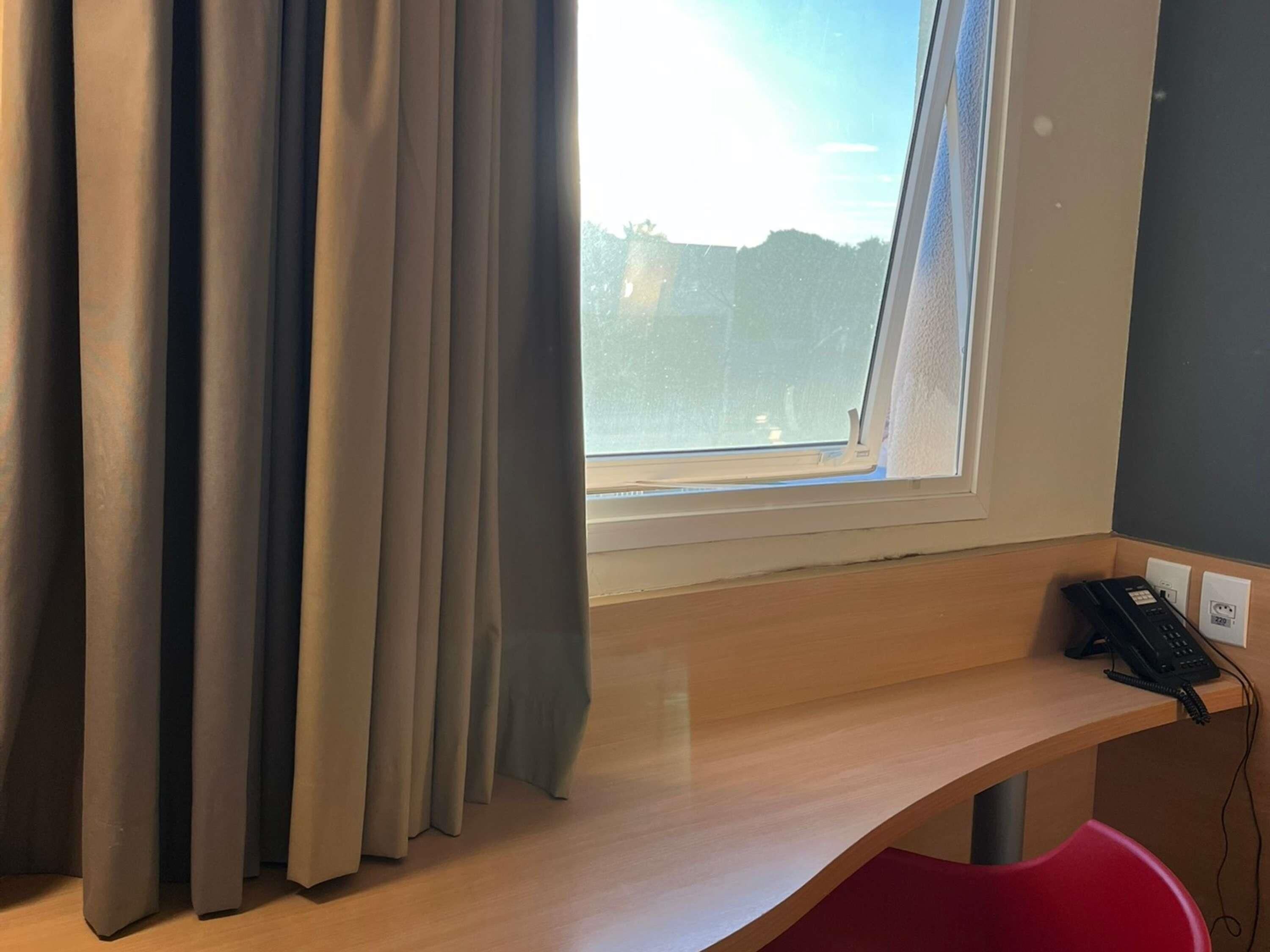 Una habitación con ventana, cortinas, un escritorio con teléfono y una silla roja.