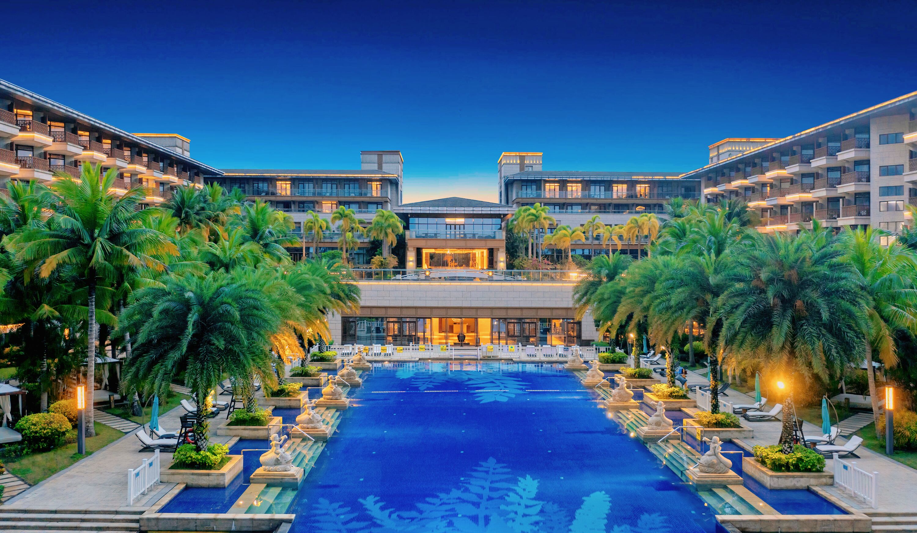 A hotel with a central pool, surrounded by palm trees and lounge chairs.