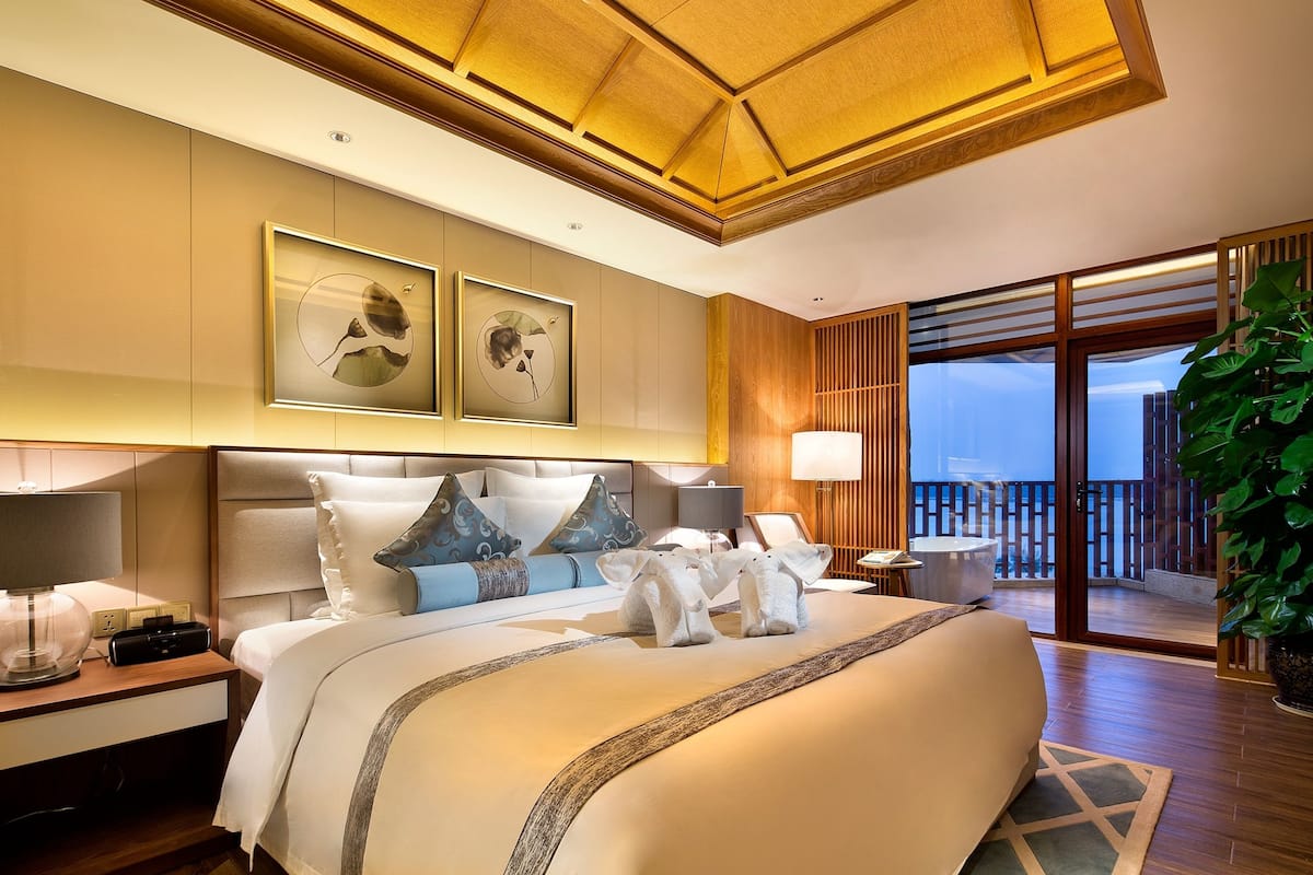 A well-lit bedroom with a large bed, two bedside tables, a nightstand, a view of the balcony, and artwork on the wall.