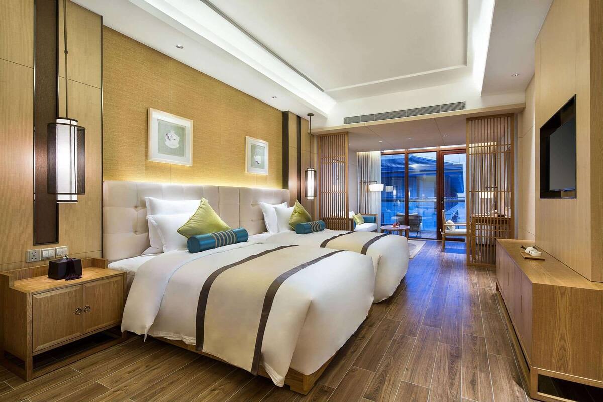 A modern hotel room with two beds, a wooden floor, a TV, and a view of the city.