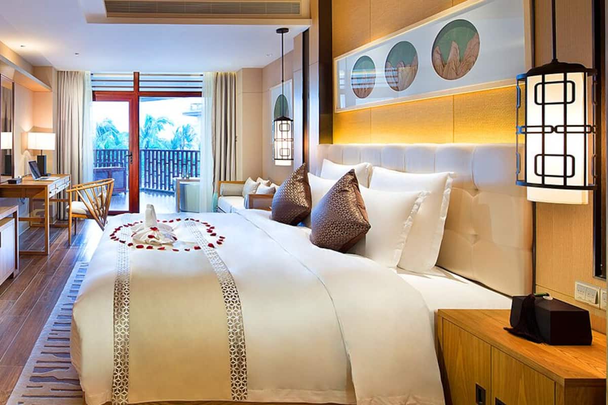 A hotel room with a large bed, a desk, a TV, and a balcony with a view of palm trees.