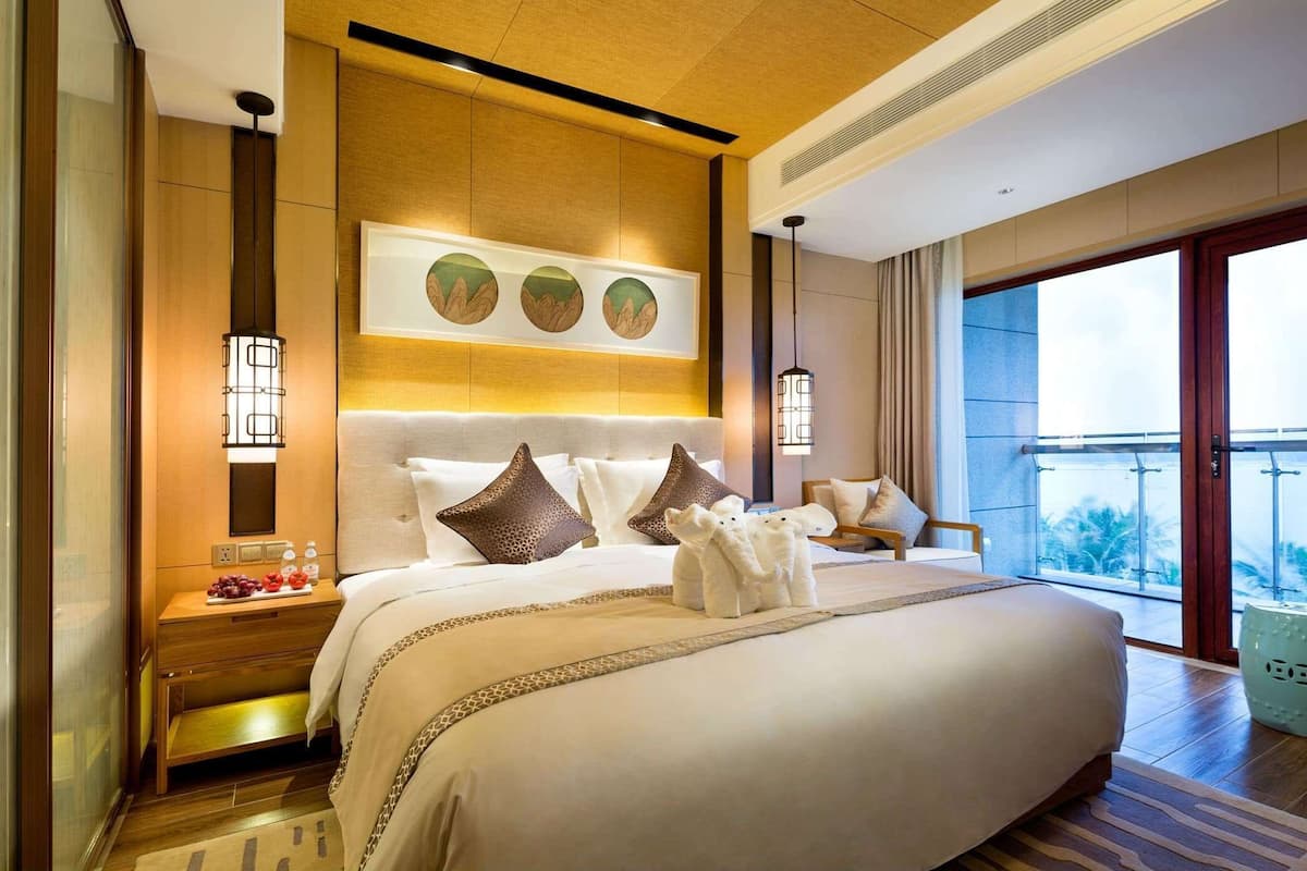 A spacious hotel room with a large bed, a nightstand with a lamp, a wall-mounted artwork, and a view of the outdoors.