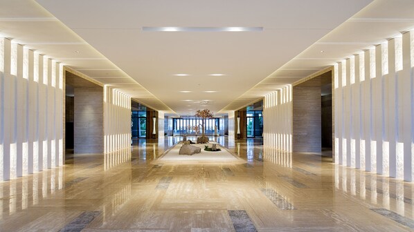 Um corredor espaçoso e moderno com piso de mármore polido, iluminação simétrica e um espaço de estar central com uma árvore e pedras.