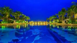 Piscina iluminada à noite, com palmeiras e guarda-sóis.