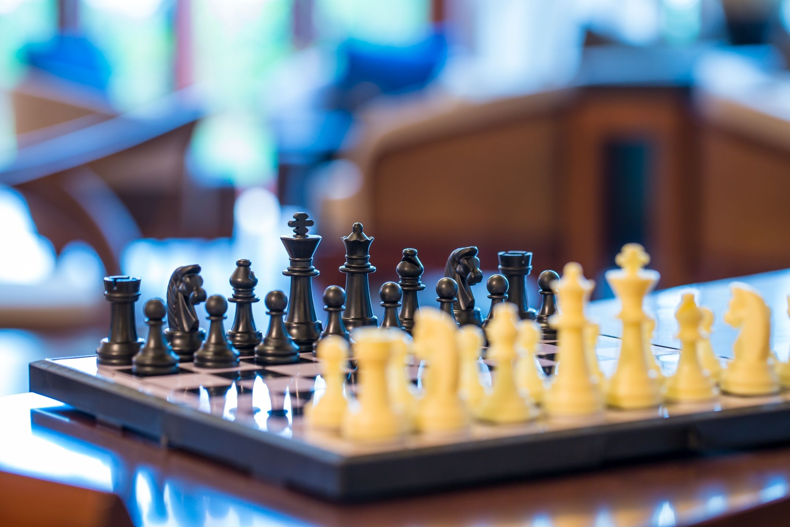 A chessboard with pieces in mid-game, set on a wooden table in a room with blurred background.