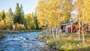 A cabin by a river with a red exterior, surrounded by trees and a clear sky. - Storsätra Fjällhotell (Idre)