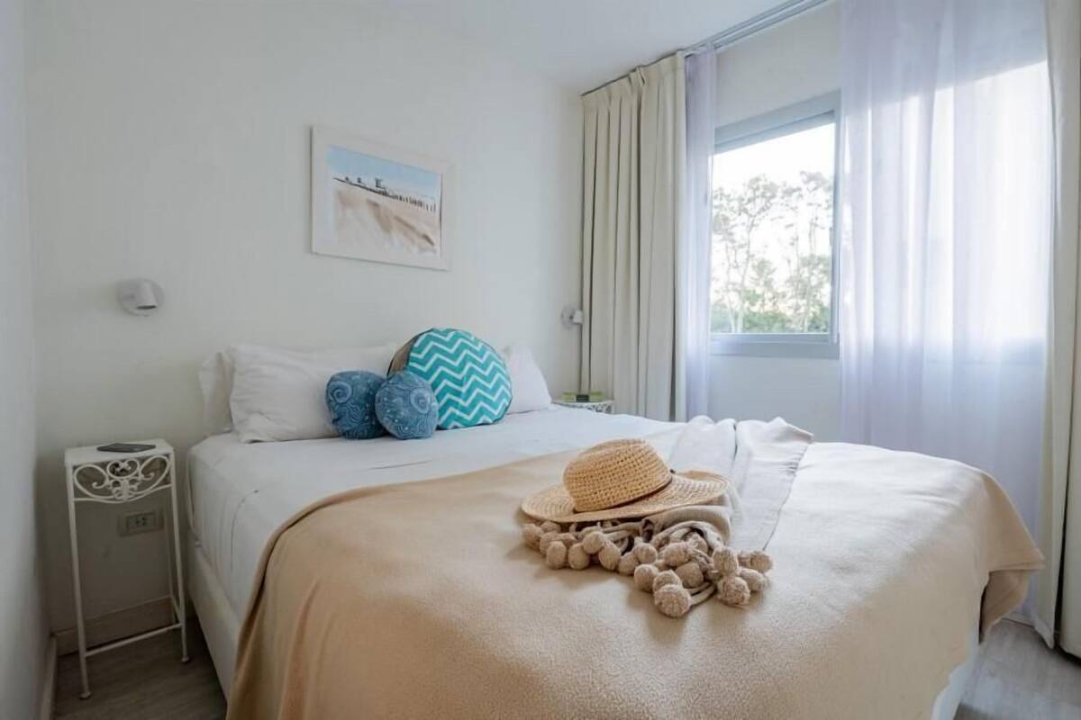 Una cama bien hecha con una manta color beige, un sombrero de paja y un cojín decorativo con borlas. Se ve una mesita de noche con una lámpara.