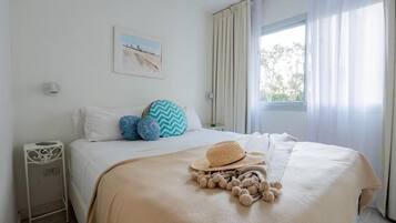 A neatly made bed with a beige blanket, a straw hat, and a decorative pom-pom throw. A small bedside table with a lamp is visible.