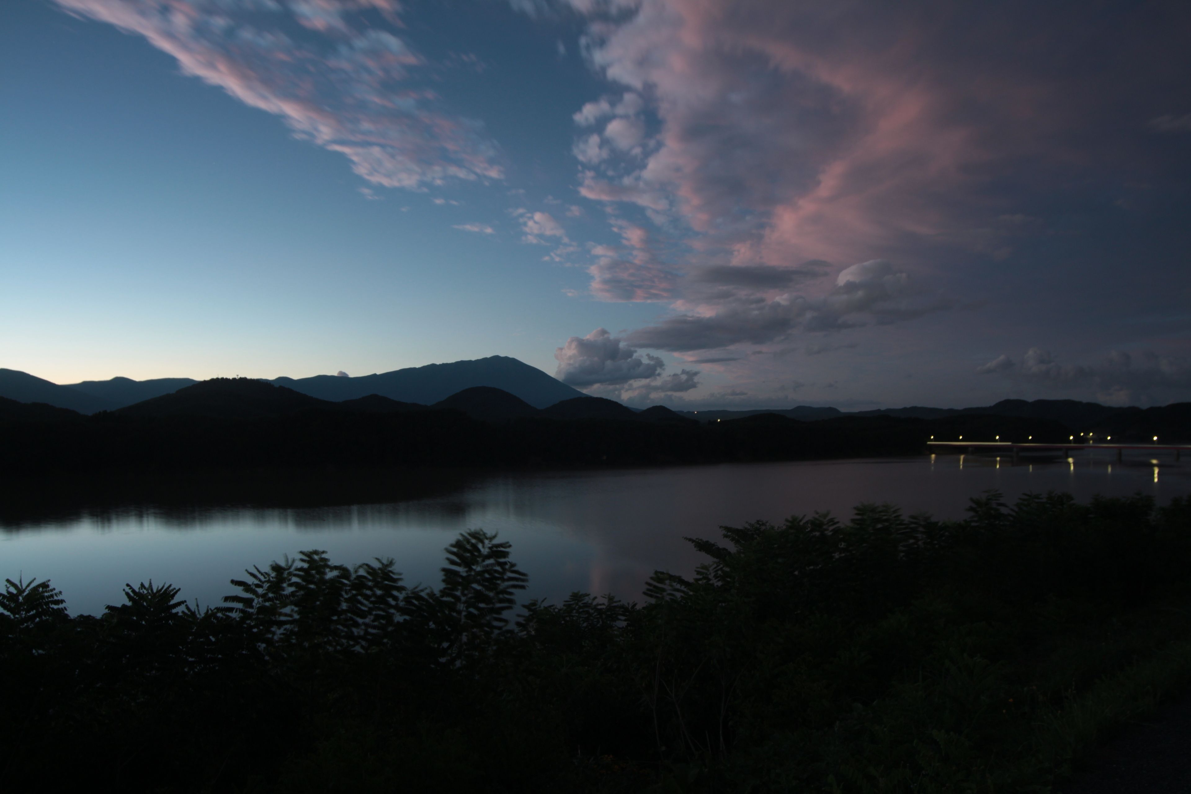 A nighttime view of a lake with mountains in the background and a bridge with lights reflecting on the water.