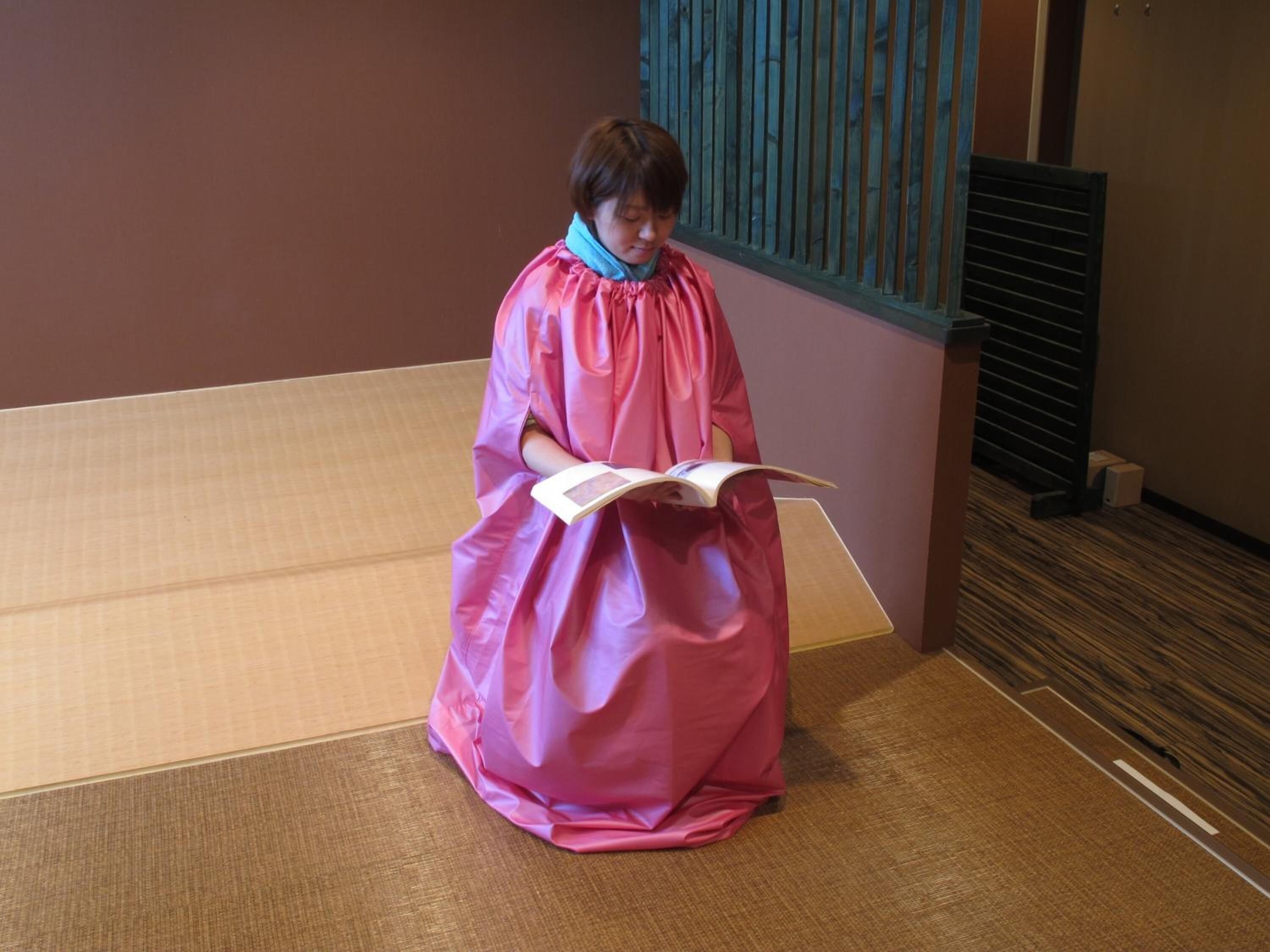 Una persona in una veste rosa seduta su un tappetino tatami in una stanza con pavimento in legno e una finestra con persiane verticali.