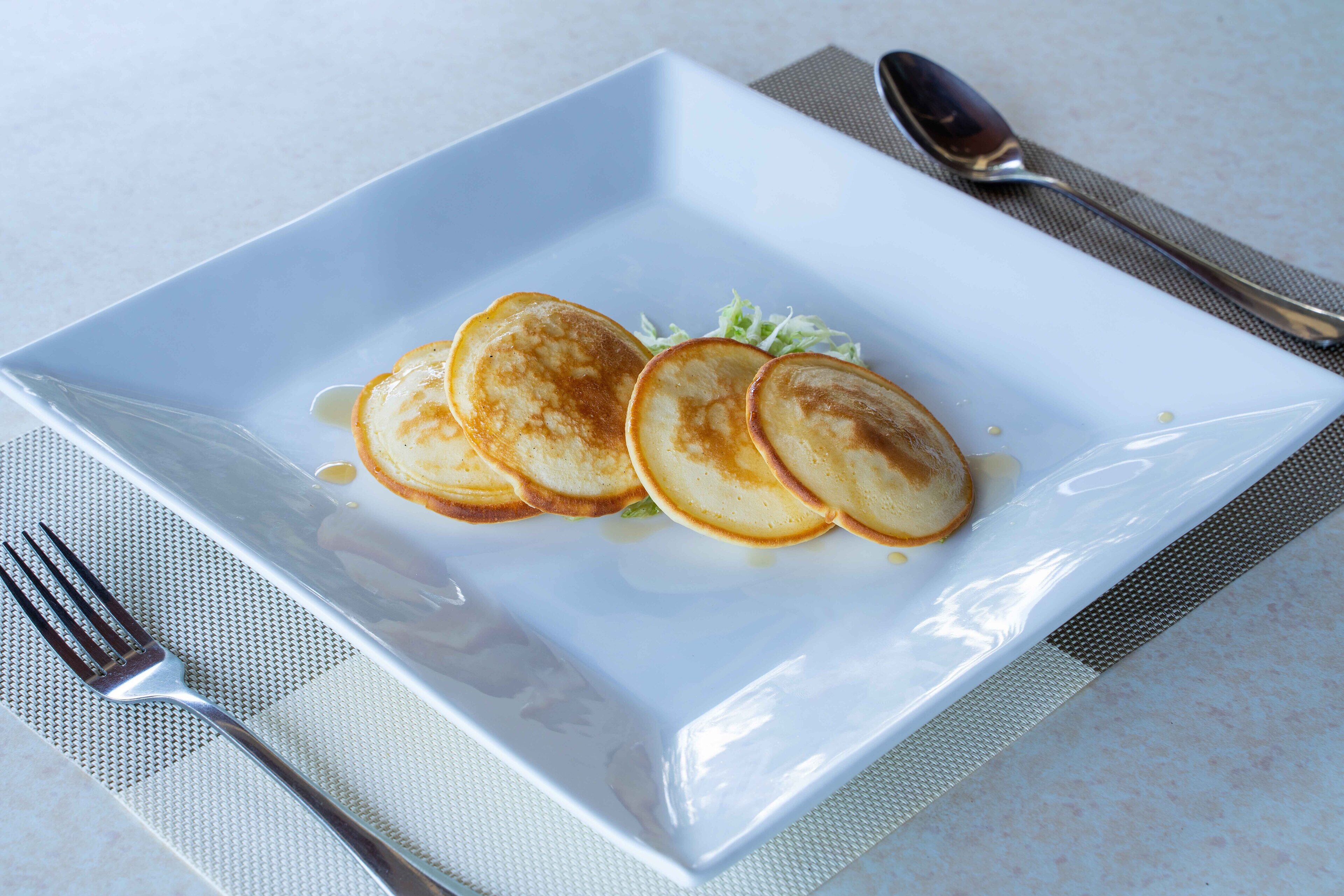 A square white plate with three pancakes and a small amount of syrup, accompanied by a fork and a spoon on a textured placemat.