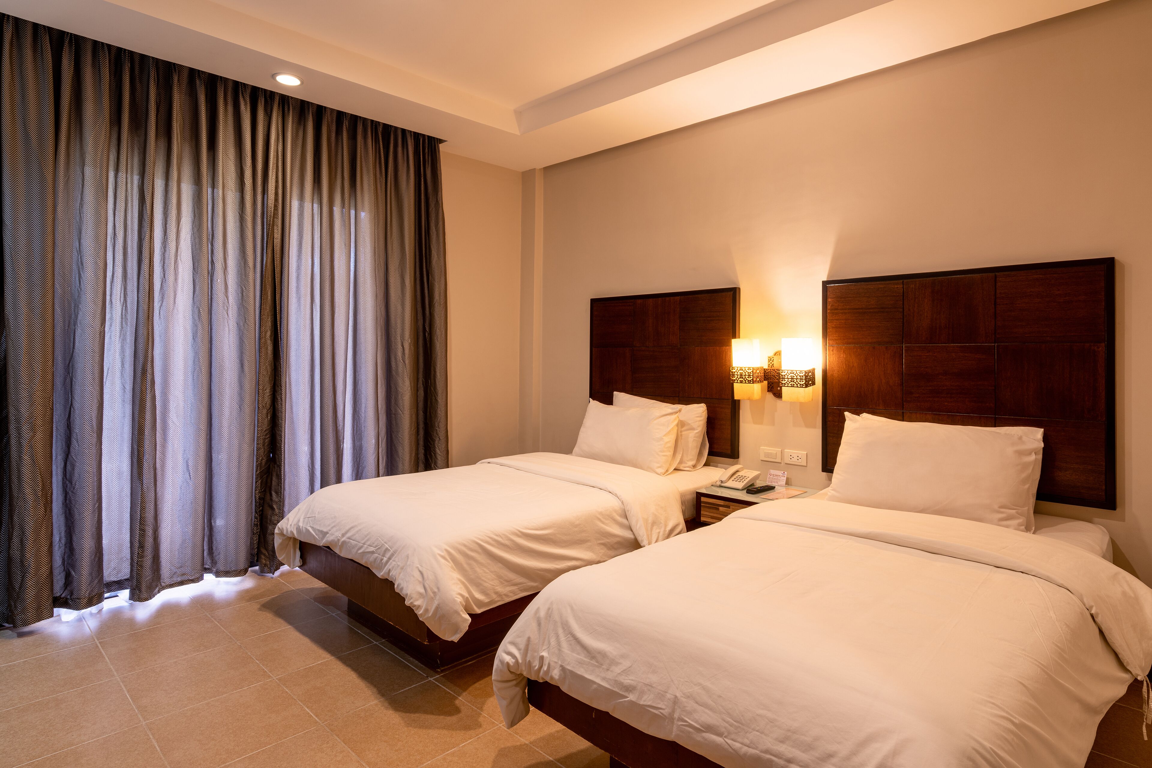 A hotel room with two beds, a large window with curtains, a wooden headboard, and a bedside table with a lamp.