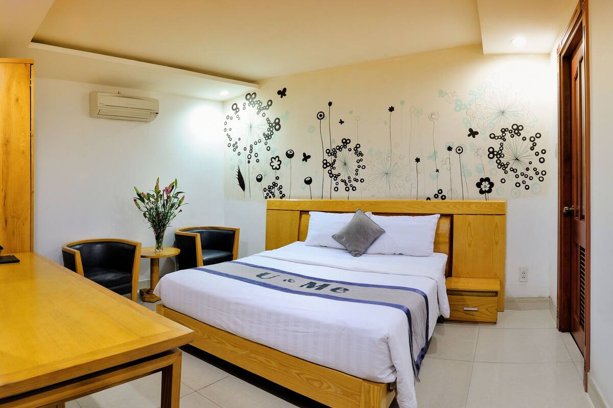 A hotel room with a wooden bed, a bedside table, a chair, a small table, and a wall decoration.
