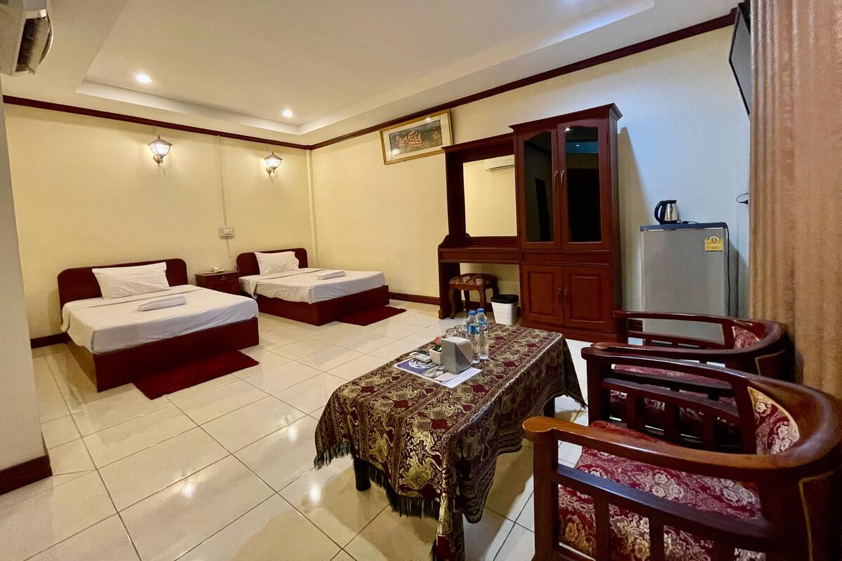 Habitación de hotel con dos camas individuales, una mesa de comedor con mantel, un armario de madera y un refrigerador.