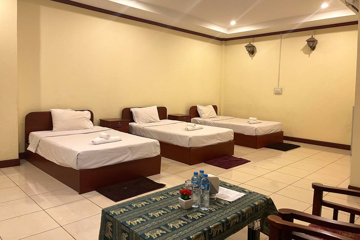 Habitación de hotel con tres camas individuales, una mesita con botellas de agua y un menú, y un tapete decorativo.
