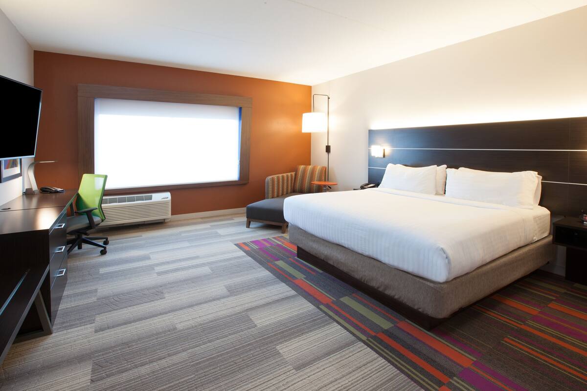Ein Hotelzimmer mit einem großen Bett, einem Schreibtisch mit Computer, einem Sessel und einem Fenster mit Jalousien.