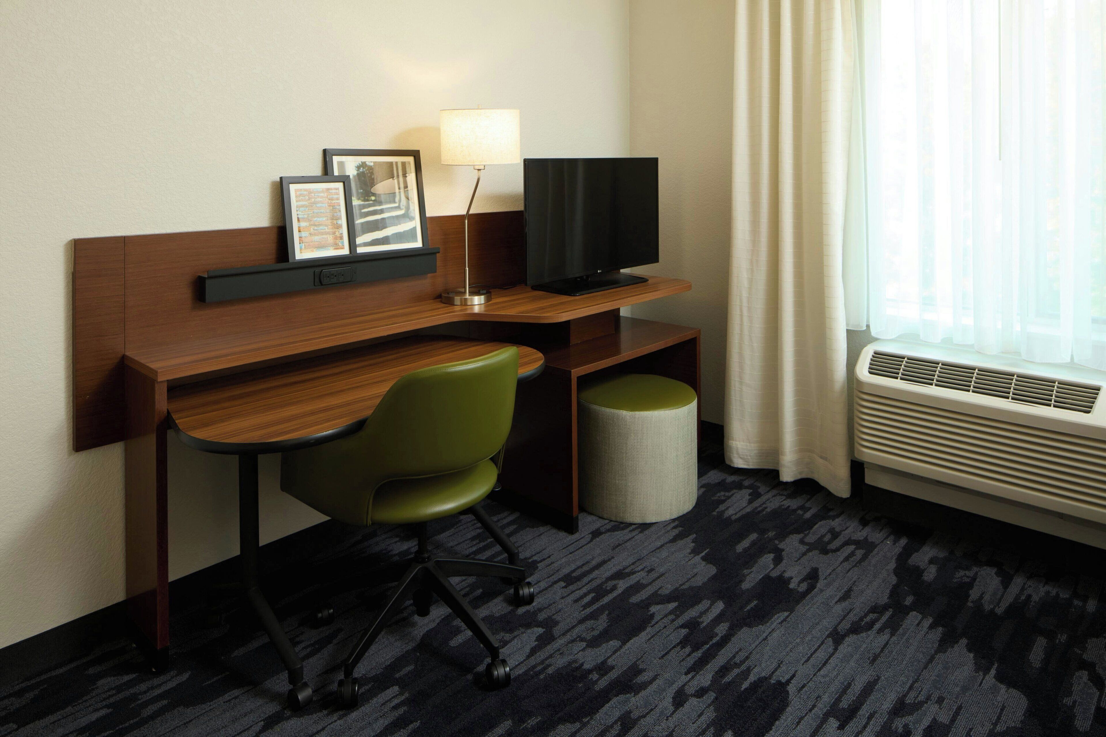 Habitación de hotel con escritorio de madera, una silla verde, televisor de pantalla plana, lámpara, cesto de basura y ventana con cortinas.
