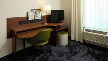 Habitación de hotel con escritorio de madera, una silla verde, televisor de pantalla plana, lámpara, cesto de basura y ventana con cortinas.
