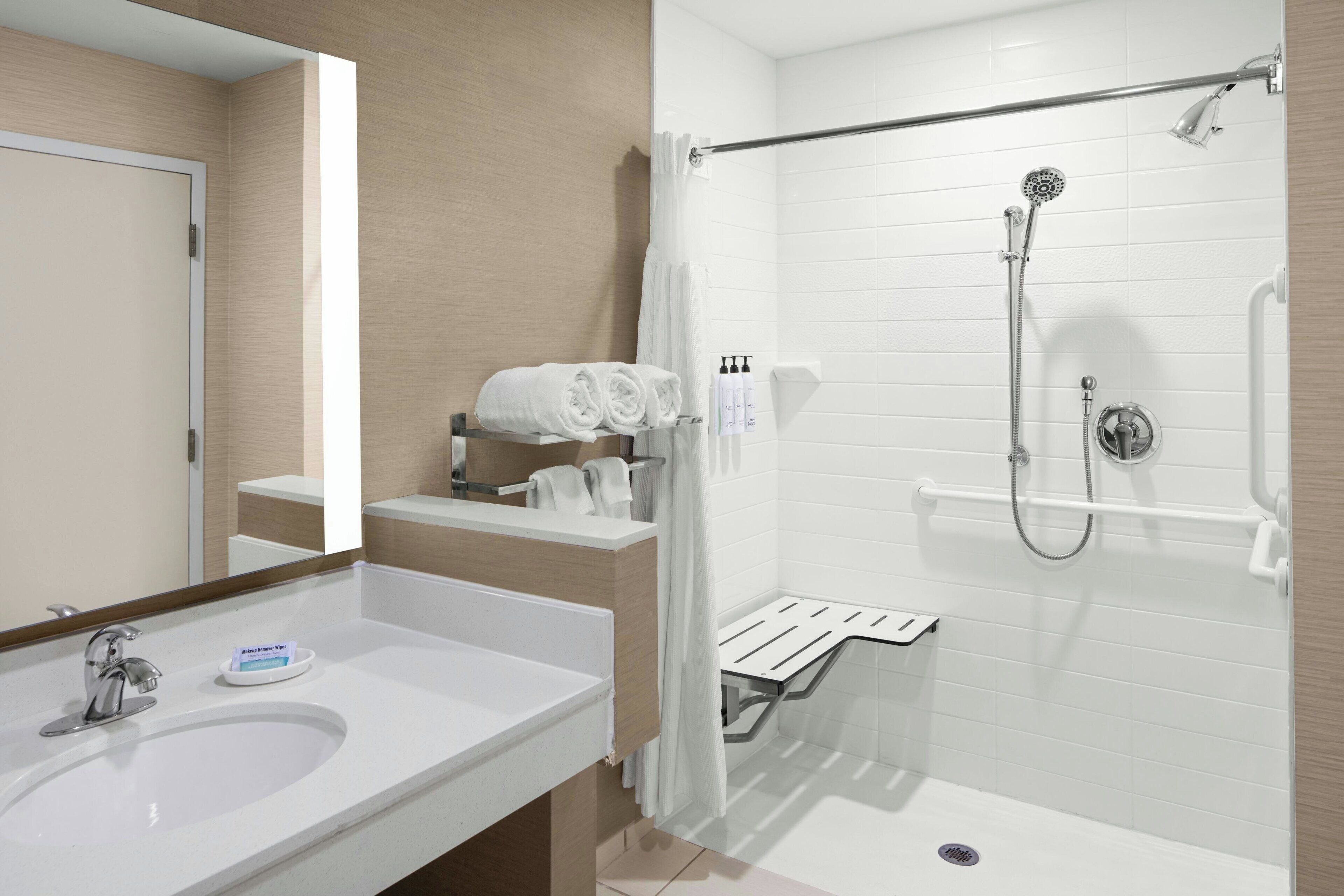 Une salle de bain moderne avec un lavabo blanc, un miroir, une douche équipée d’un pommeau de douche à main et des serviettes soigneusement pliées, rangées sur une étagère.