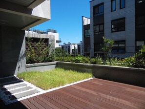 Una terraza moderna al aire libre con cubierta de madera, macetas de hormigón con plantas y vistas a edificios cercanos.