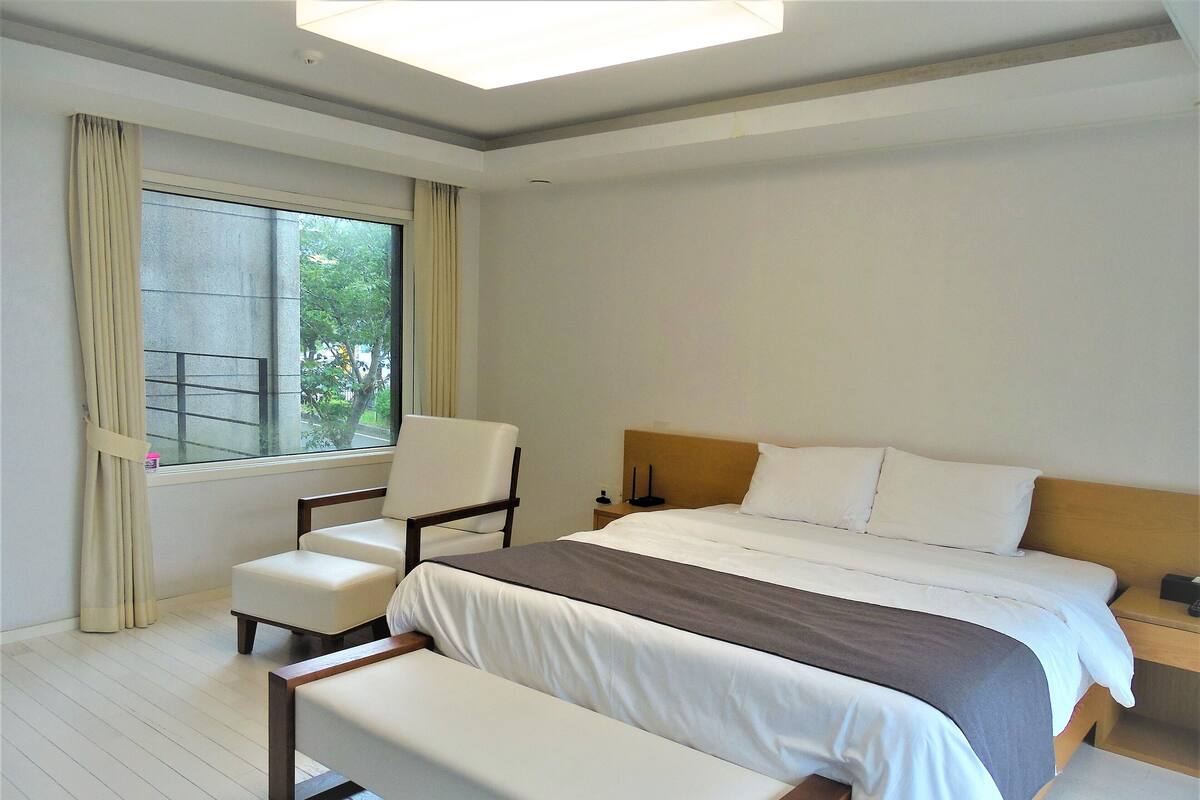 Une chambre à coucher comprenant un lit, une chaise, un banc, une table de chevet et une fenêtre avec des rideaux.