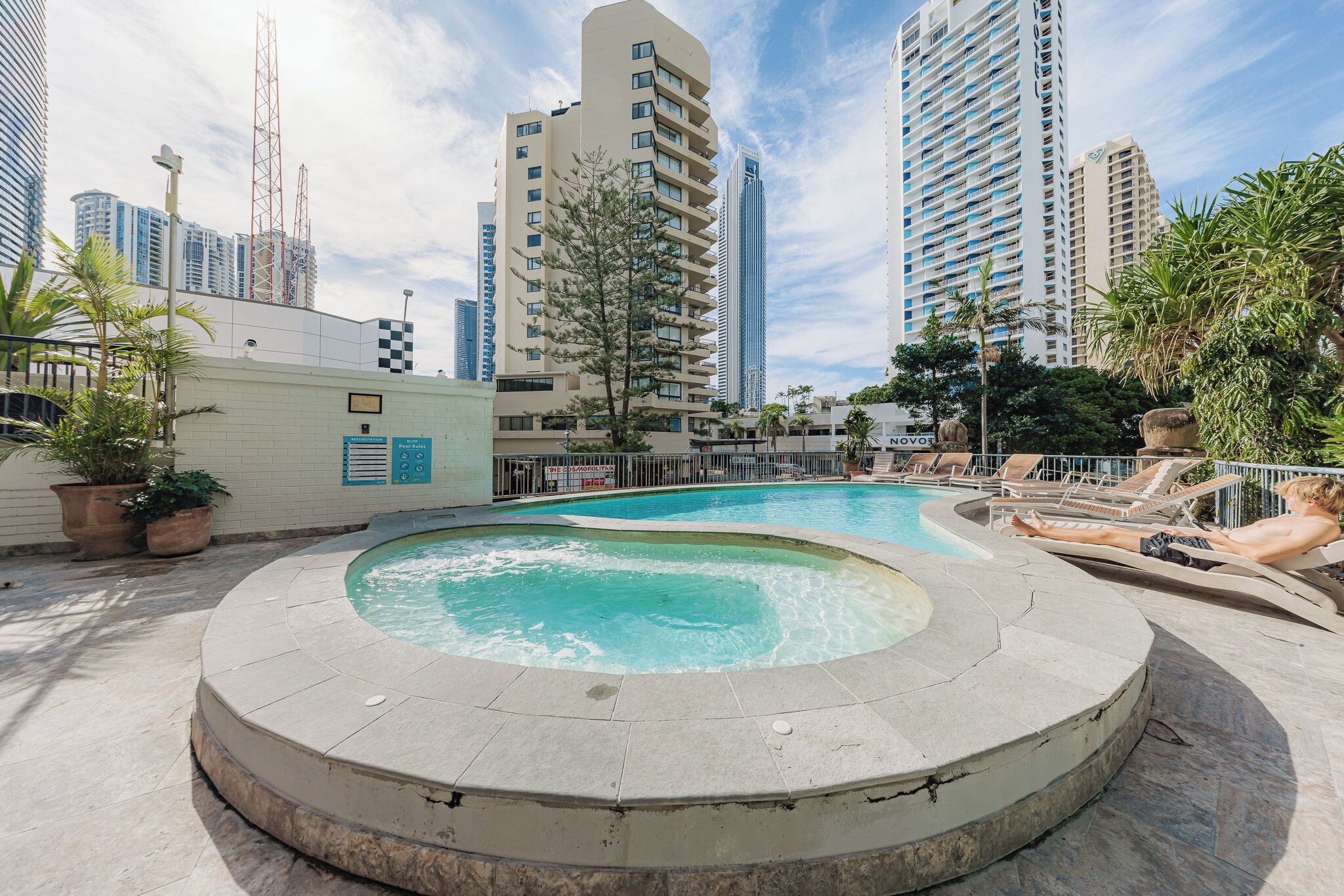 Una piscina en la azotea con un jacuzzi, rodeada de edificios altos y palmeras.