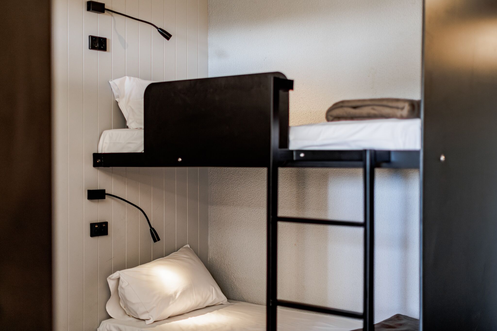 Ein Etagenbett mit Kissen und Decke auf der oberen Koje, eine Lampe seitlich und eine Steckdose an der Wand.