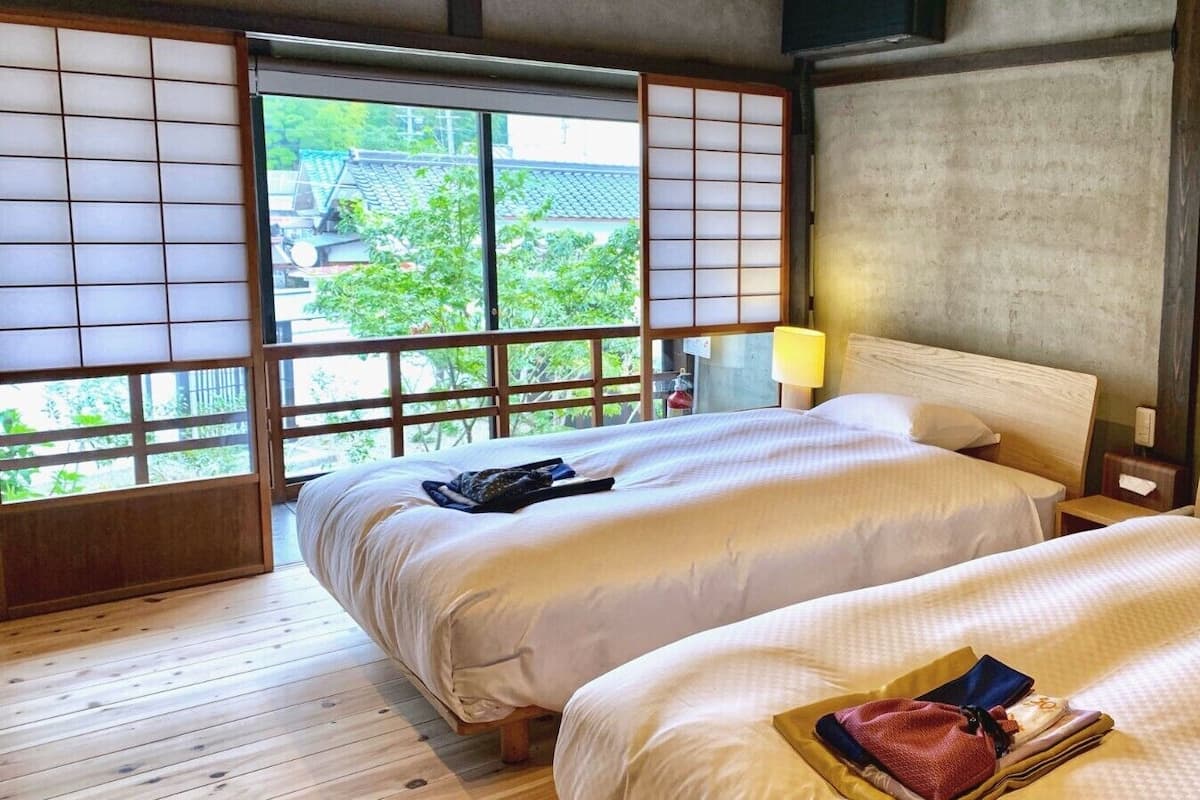 Una habitación tradicional japonesa con suelo de tatami, dos camas con ropa blanca, cabeceros de madera y vistas a la vegetación a través de biombos shoji.