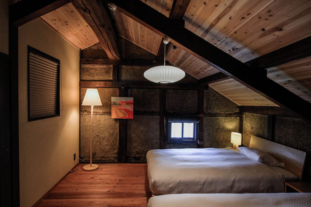 Un dormitorio acogedor en el ático con vigas de madera a la vista, una cama grande, una lámpara de pie y un ventanal pequeño.