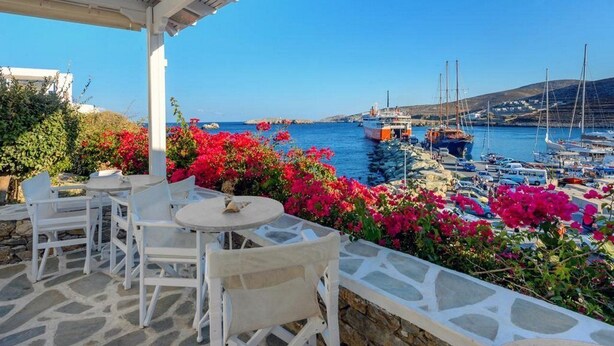 Une terrasse en bord de mer, agrémentée de meubles blancs et de fleurs aux couleurs vives, qui offre une vue sur une marina où sont amarrés des bateaux, le tout sous un ciel bleu azur.