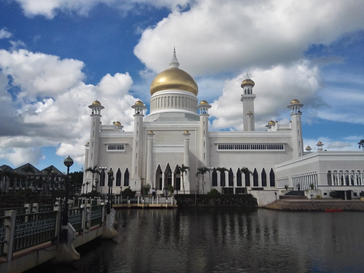 Un grand bâtiment blanc surmonté d’un dôme doré et flanqué de plusieurs minarets, entouré d’eau et de palmiers.