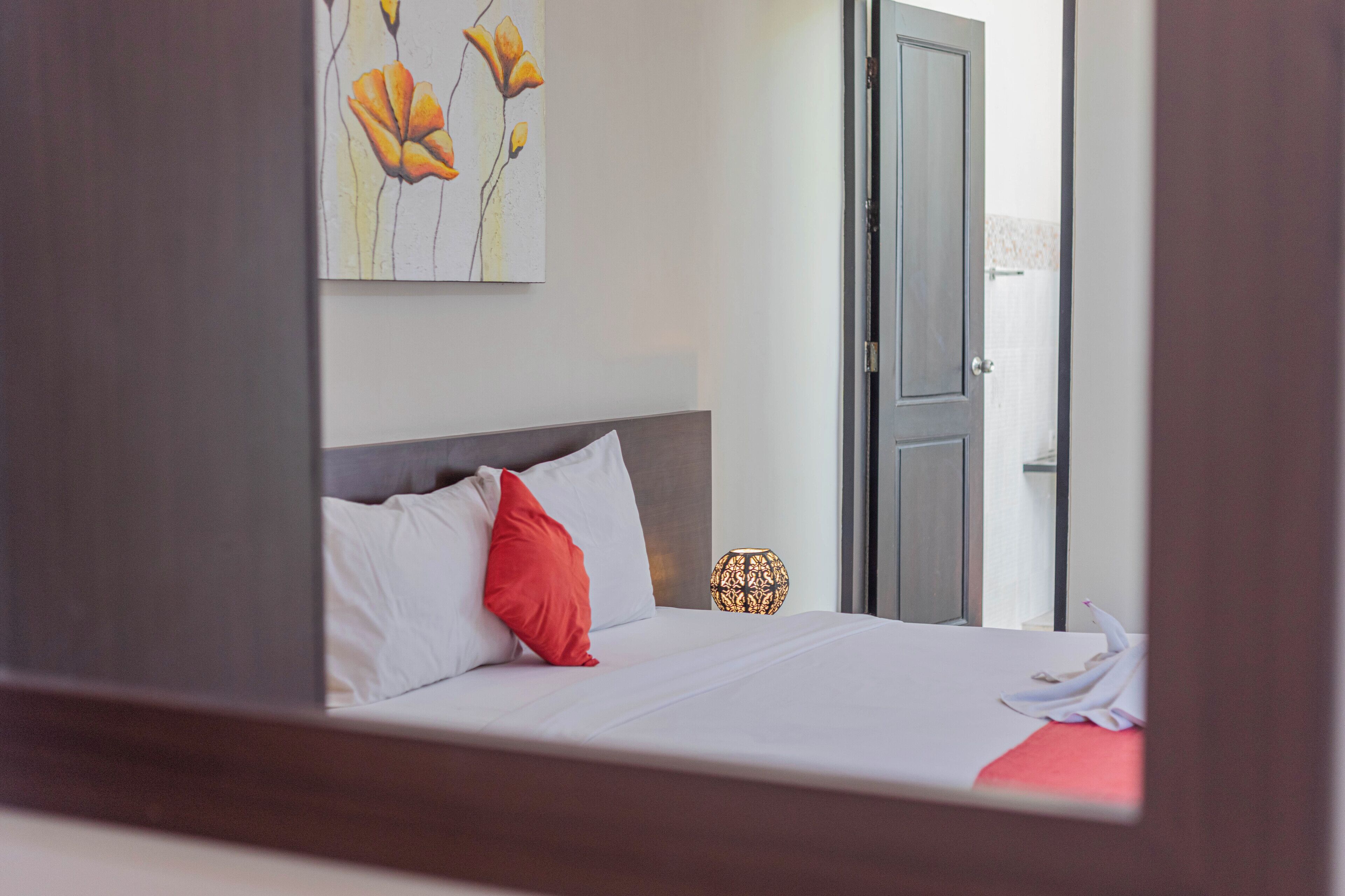 A neatly made bed with white and red pillows, a decorative wall piece, and a small table with a patterned lamp.