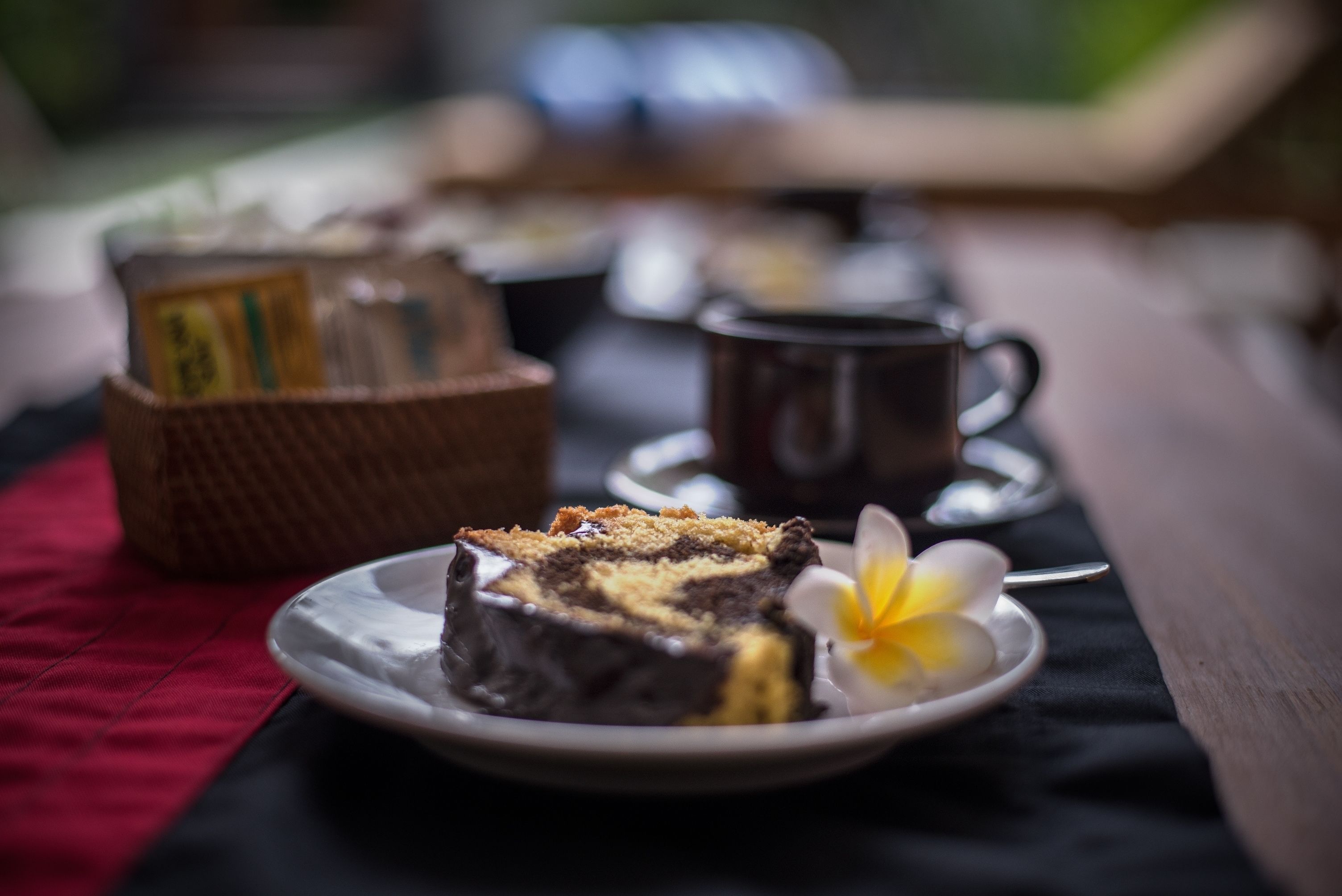 A slice of cake on a plate with a flower, a cup, and a basket of tea on a table.