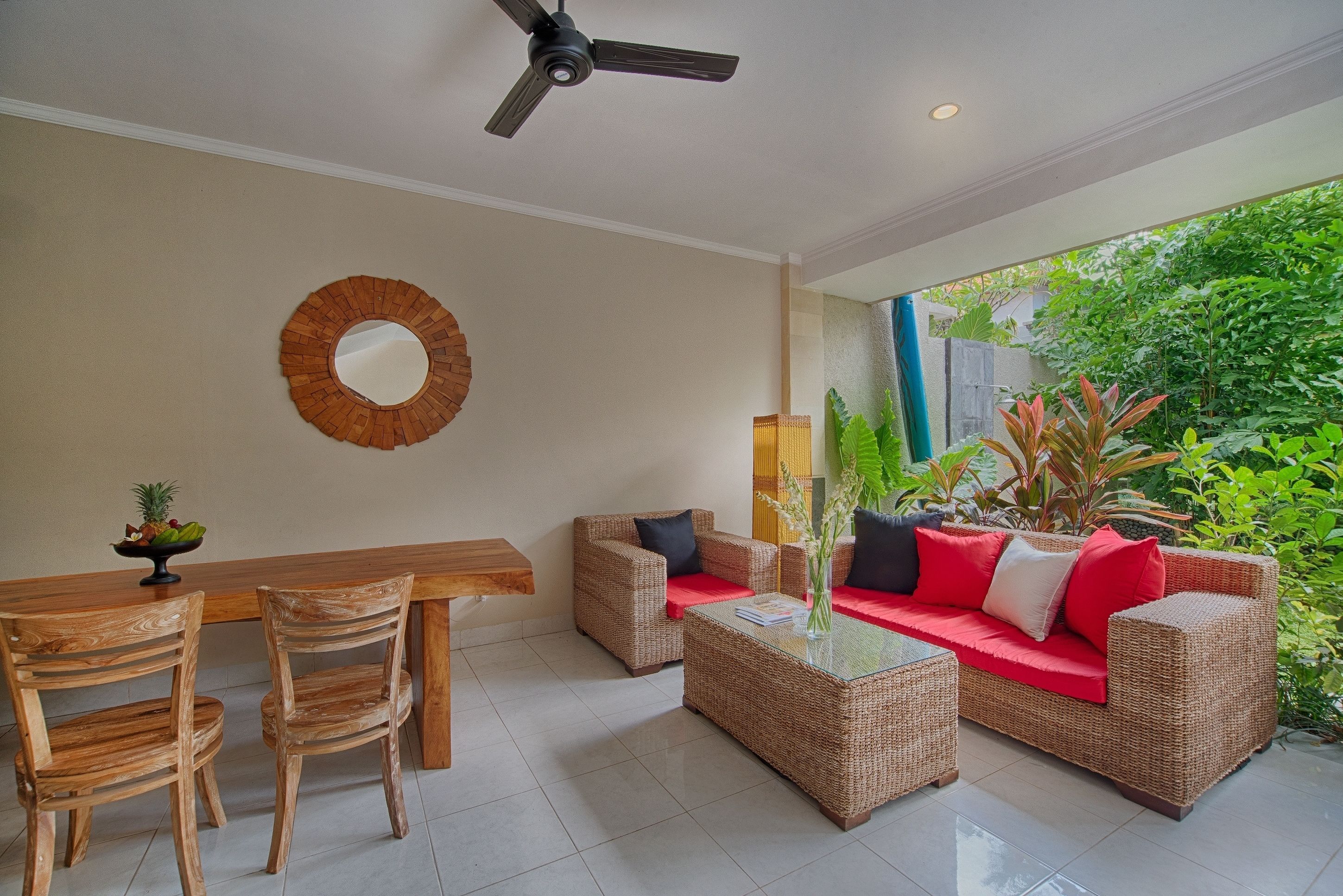 A room with a ceiling fan, a round mirror, wicker furniture, and a view of greenery outside.