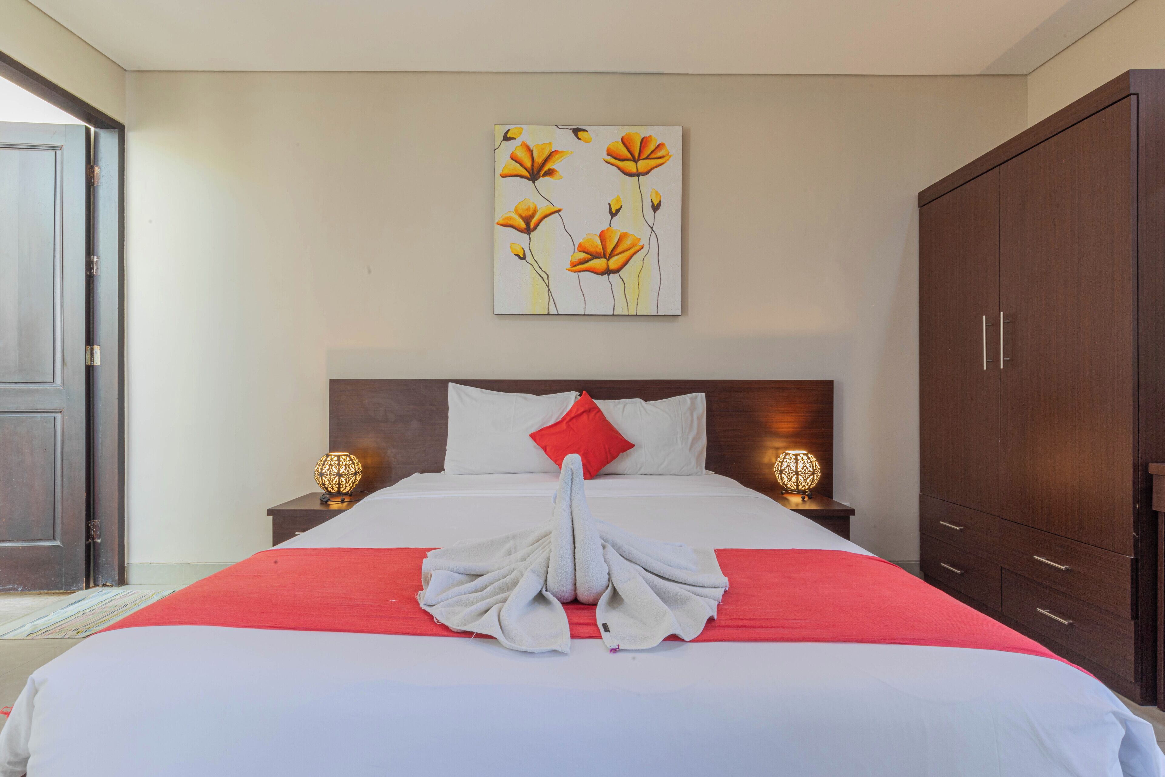 A neatly made bed with a red blanket and a towel arranged in a swan shape, a white pillow, and a red pillow on a wooden headboard. There is a painting of flowers on the wall above the bed, and two bedside lamps on wooden nightstands.