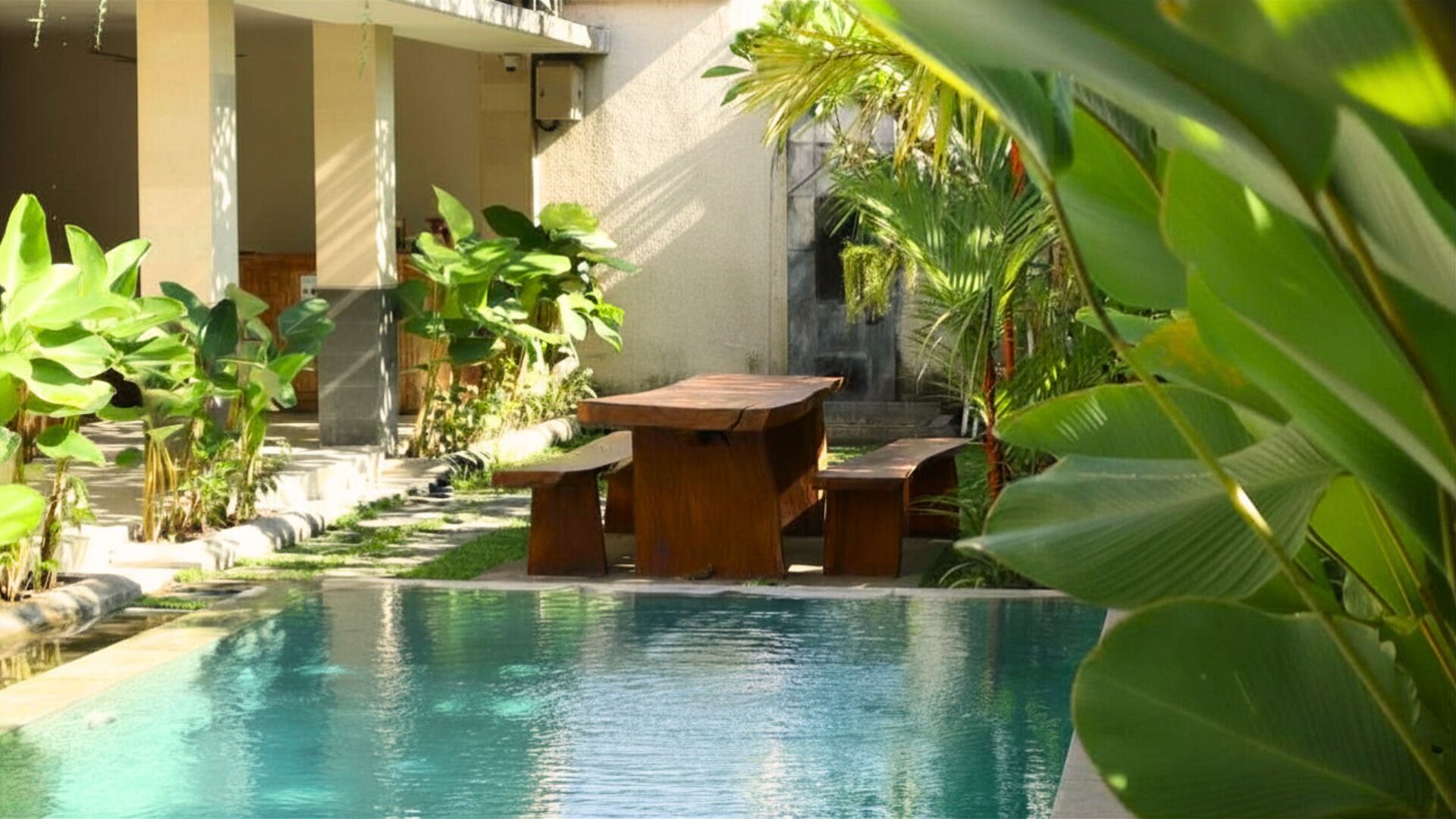 Un espace piscine agrémenté d’une table et de chaises en bois, entouré d’une végétation luxuriante et d’un mur en pierre.