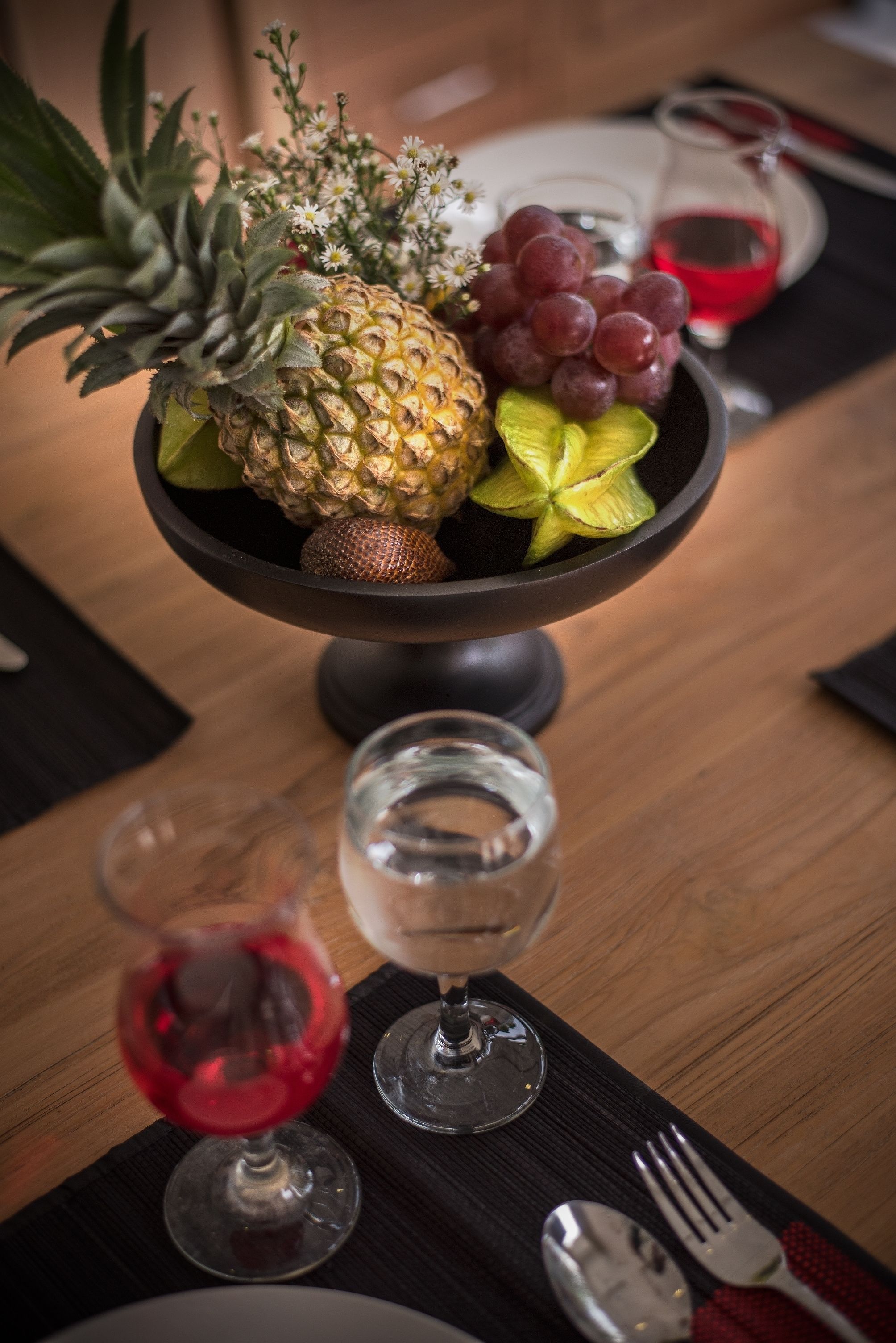 A dining table with a fruit bowl containing pineapple, grapes, and a starfruit, accompanied by two wine glasses and a red wine bottle.