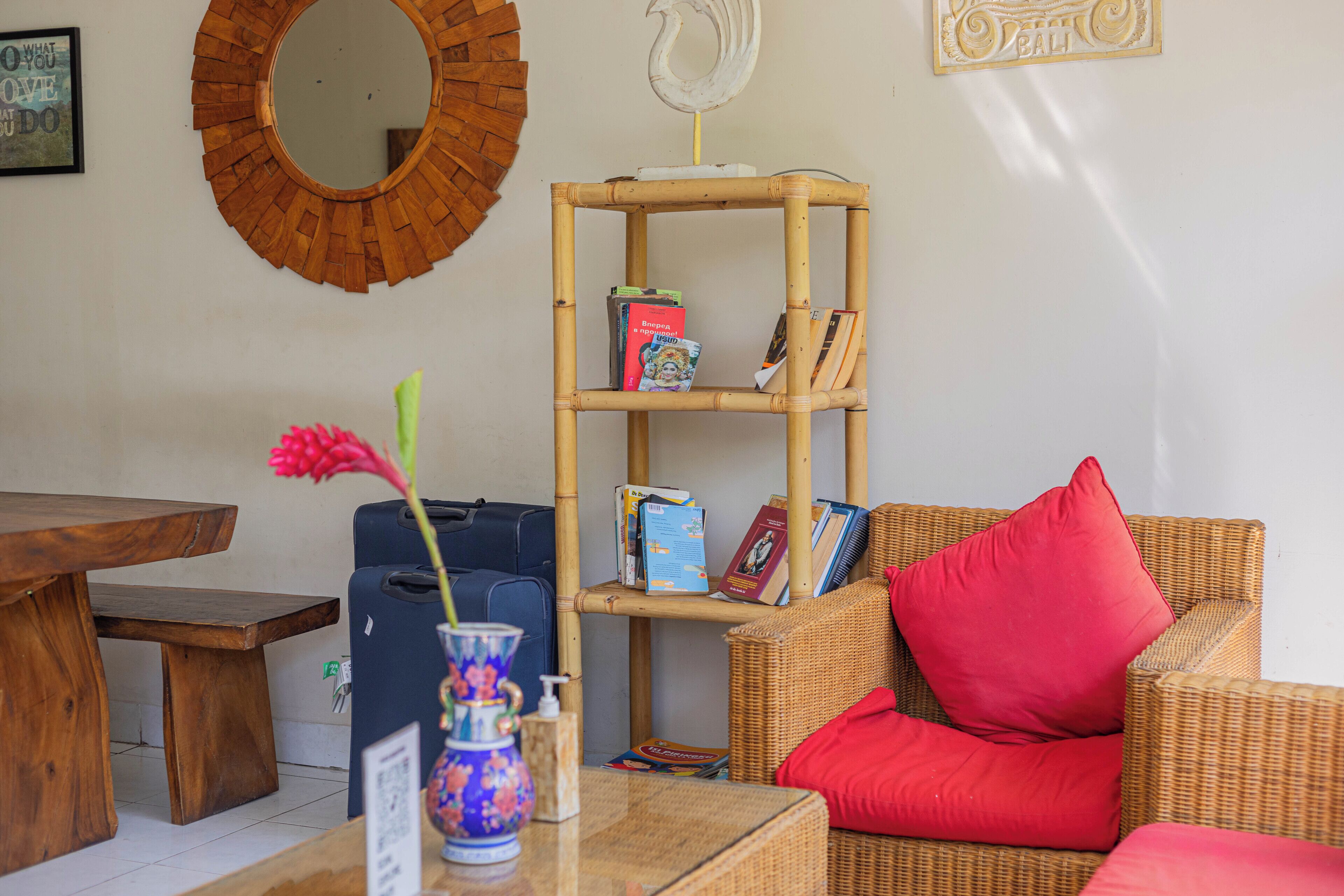 A room with a wooden table, a wicker sofa with red cushions, a bamboo bookshelf, and a blue suitcase.