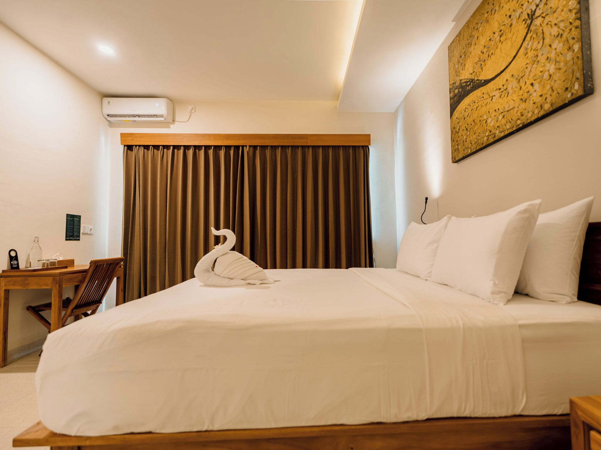 Ein Hotelzimmer mit einem großen Bett, einem Holztisch, einem Stuhl, einem schwanenförmigen Kopfkissen und einer Wand-Klimaanlage.