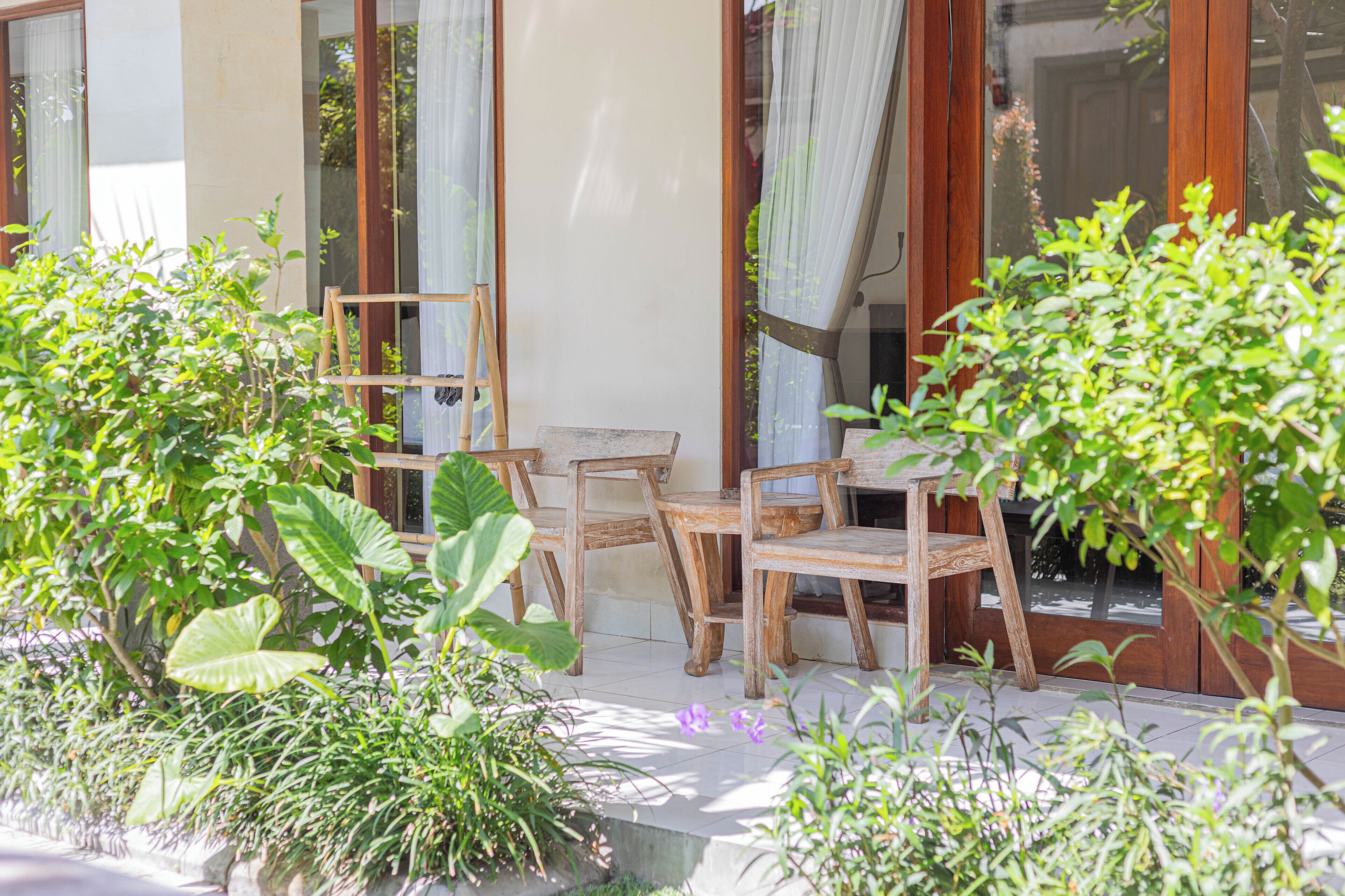 Een terras met houten stoelen en een tafel, omgeven door weelderige begroeiing en een groot raam met gordijnen.