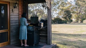 A person cooking outdoors on a grill, with a wooden house and a grassy yard in the background. - GRAMPIANS HISTORIC TOBACCO KILN (Moutajup)