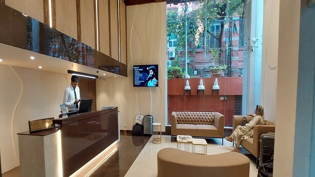 A modern hotel lobby with a reception desk, a large flat-screen TV, a comfortable seating area, and a view of the outside. - Lytton Hotel (Kolkata)