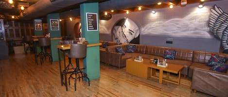A cozy restaurant interior with wooden floors, a large mural on the wall, and a seating area with a sofa and bar stools.