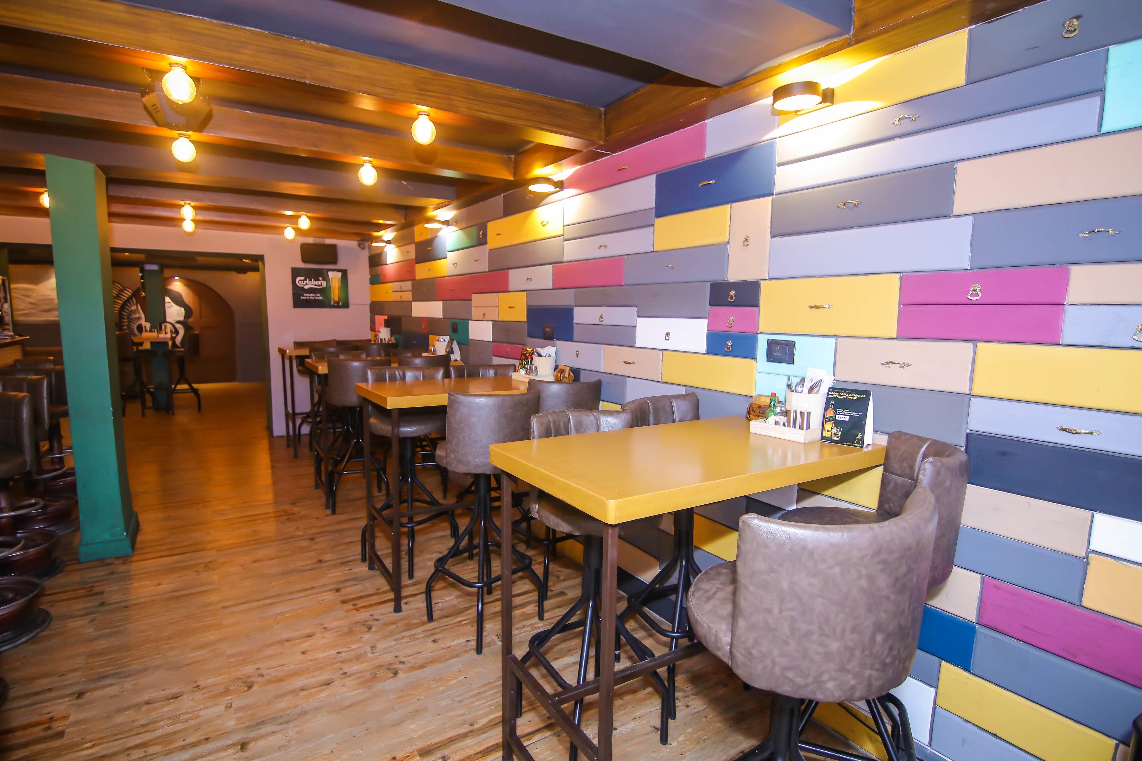 Un interior de restaurante con suelos de madera, varias mesas y sillas, y una pared colorida con cajones.