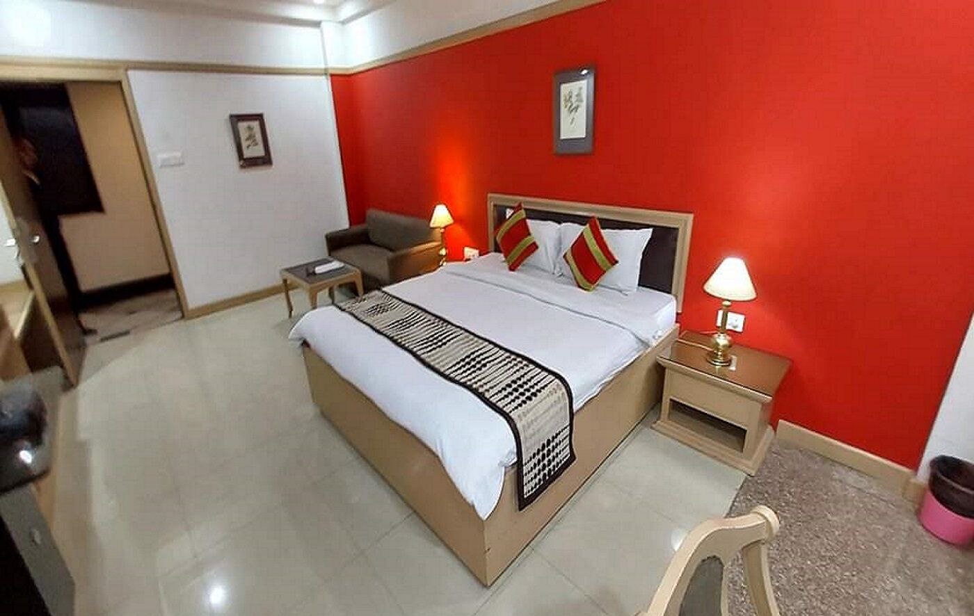 A hotel room with a bed, bedside tables, a nightstand, a chair, and a red wall.
