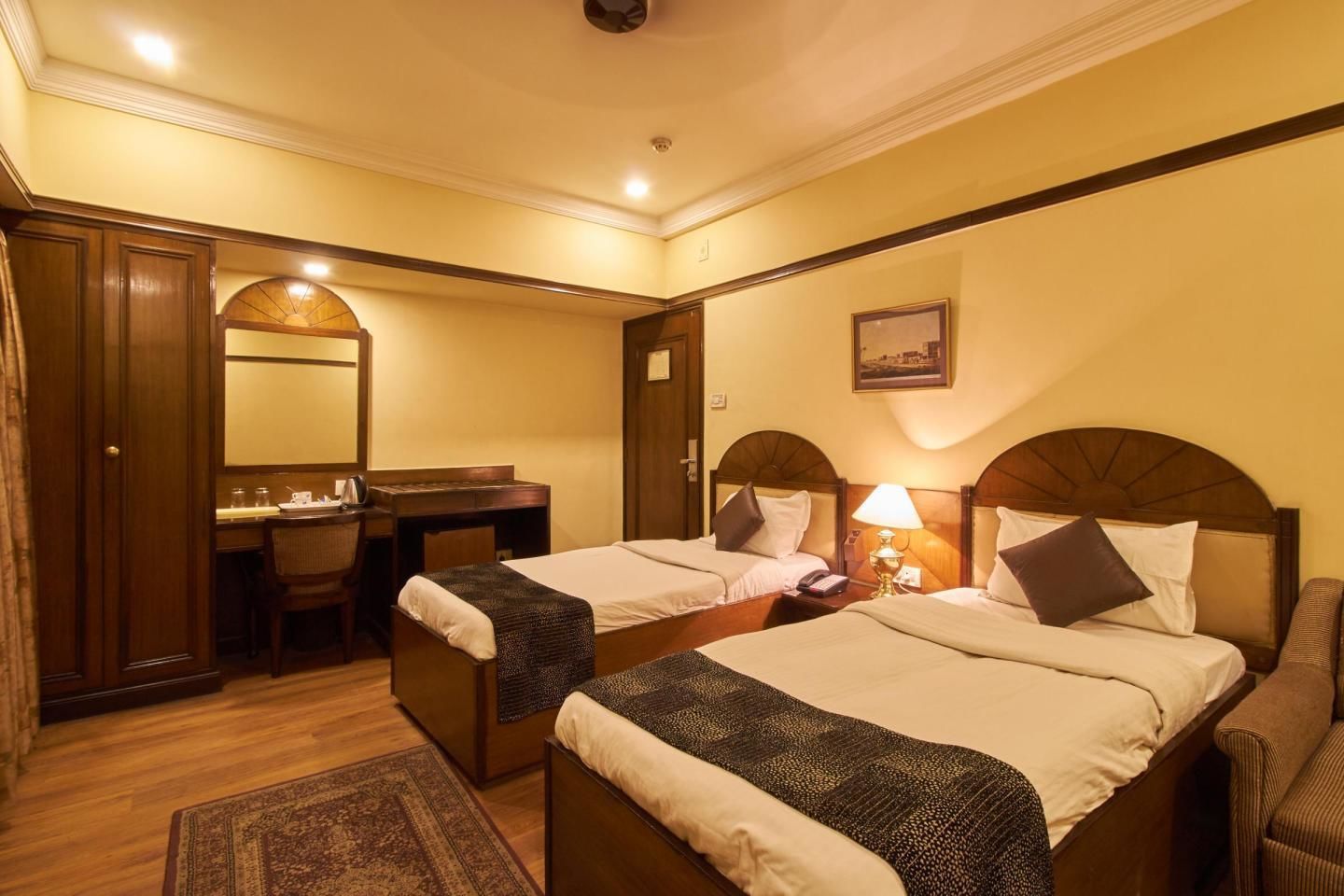 A hotel room with two single beds, a desk, a chair, a mirror, and a small rug.